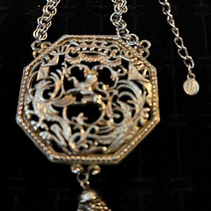 May include: A silver necklace with a large octagonal pendant featuring an intricate filigree design. The pendant has a tassel hanging from the bottom.