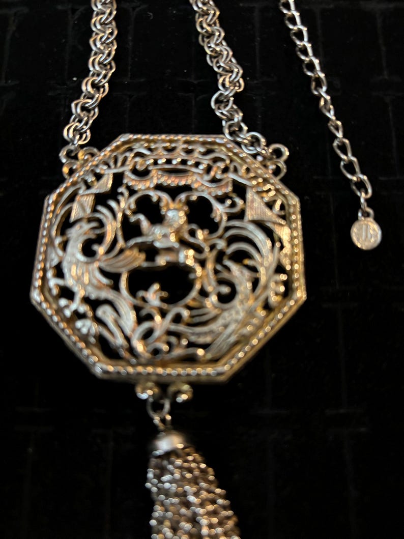 May include: A silver necklace with a large octagonal pendant featuring an intricate filigree design. The pendant has a tassel hanging from the bottom.