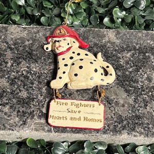 Può includere: Un ornamento a forma di cane dalmata con un cappello da pompiere rosso con il numero 1. Il cane ha macchie nere e un collare rosso. Un cartello recita "Fire Fighters Save Hearts and Homes".