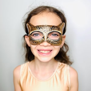 May include: A young person wearing a gold and black glitter cheetah mask with a light brown tank top.