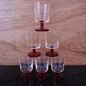 Op de afbeelding: Een set van zes heldere glazen bekers gerangschikt in een piramidevorm. Elke beker heeft een cilindrische kom en een korte, stevige steel verbonden met een ronde voet, allemaal vervaardigd uit dieprood glas. De glazen zijn transparant.