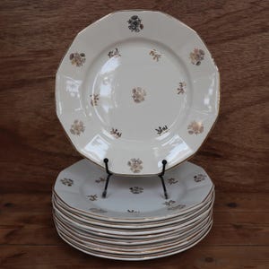 Peut inclure: Une pile de 10 assiettes plates en céramique blanche avec un bord doré et un motif floral. Les assiettes sont empilées sur une surface en bois.
