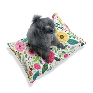 Può includere: Un piccolo cane grigio sdraiato su un letto per cani bianco con motivo floreale. Il letto ha fiori rosa, gialli e verdi.