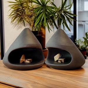 May include: Two dark gray, teardrop-shaped ceramic fireplaces with open fronts, each containing pieces of firewood. The fireplaces are displayed on a wooden surface, with green plants in the background. A modern home decor item.
