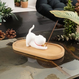 Könnte beinhalten: Weißer Keramikkätzchen-Räucherhalter mit rotem Halsband und einer goldenen Glocke. Die Katze liegt auf der Seite mit dem Kopf zur Seite gedreht. Der Räucherhalter steht auf einem Holztablett.