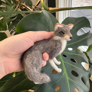 May include: A gray and white needle felted wolf figurine with brown ears. The figurine is sitting on a green leaf.