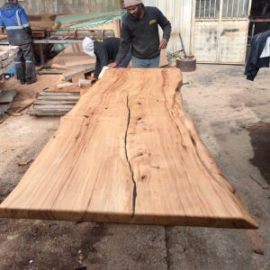 Peut inclure: Une grande table en bois non finie avec un bord naturel et une fissure importante au centre. Le bois est de couleur brun clair avec des motifs de grain visibles. La table est dans un atelier.