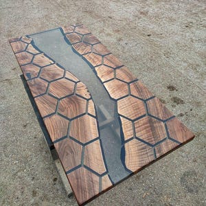Op de afbeelding: Een houten tafelblad met een doorlopende rivier van heldere epoxyhars. Het hout is donkerbruin met een honingraatpatroon.
