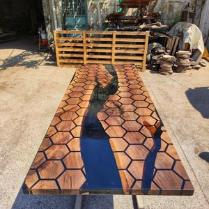 Puede incluir: Una mesa de madera rectangular grande con un dise&ntilde;o &uacute;nico. La superficie presenta un patr&oacute;n de panal con resina oscura que rellena los espacios. Un r&iacute;o de resina azul oscuro recorre el centro. La mesa est&aacute; soportada por patas de metal.