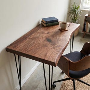 May include: A wooden desk with a live edge and black hairpin legs. The desk has a natural wood grain finish. Two books and a mug on a coaster sit on the desk. A wooden and black chair is next to the desk.
