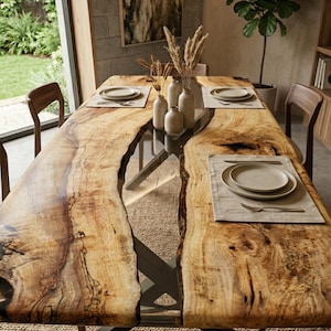 May include: A large, rectangular wooden dining table with a live edge design. The table features a natural wood grain pattern with a clear epoxy resin center. Set with neutral-toned place settings, the table is supported by a dark metal base.