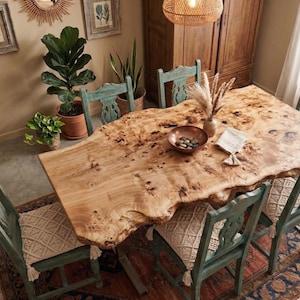 Könnte beinhalten: Ein großer, hellfarbiger Esstisch aus Holz mit einer Baumkante, umgeben von sechs teal-farbenen Stühlen mit gewebten Sitzkissen. Der Tisch ist mit einer Kupferschale, einer Vase und dekorativen Gegenständen gedeckt. Der Raum hat einen Teppich und Pflanzen.