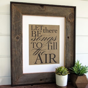 May include: A rustic wood framed print with the text "LET there BE songs TO fill the AIR" on a burlap background. The print is sitting on a wooden surface with two small potted plants.