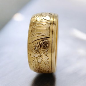 Pode incluir: Um anel dourado com gravuras detalhadas. O anel apresenta um brasão de armas e desenhos de folhas de carvalho e bolotas. A banda do anel é espessa e tem um acabamento fosco, mostrando as intrincadas esculturas.