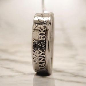 May include: Close-up of a silver coin from 1924. The coin has the word "DANMARK" vertically engraved on its side. The coin's edge is detailed with the year and other markings. The coin is set against a blurred background.