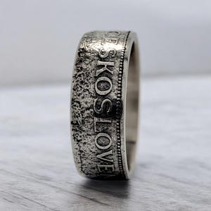 Puede incluir: Un anillo de plata hecho con una moneda de la República Checa. El anillo tiene la inscripción "CESKOSLOVENSKO" alrededor de la banda.