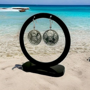 May include: Silver coin earrings with a five-cent design, displayed on a black stand against a beach backdrop. The earrings are round with a detailed emblem and are suspended from silver hooks. The ocean and sand create a scenic background.