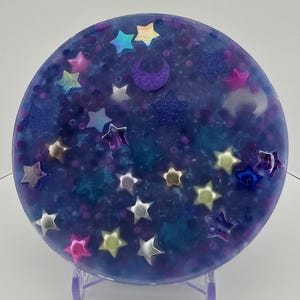 May include: A round, blue resin coaster with embedded stars in various colors, including silver, pink, and iridescent. A purple crescent moon and a glittery star are also featured. The coaster is displayed on a clear stand.