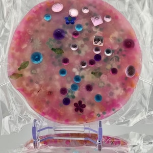 May include: A round, pink resin coaster with embedded colorful beads, gems, and floral accents. The coaster is displayed on a clear plastic stand, showcasing its decorative design. The coaster is a unique, handmade item.