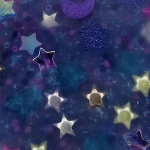 K&ouml;nnte beinhalten: Nahaufnahme eines blauen, glitzernden Harzornaments mit eingebetteten Sternen und einem Halbmond. Die Sterne sind in verschiedenen Farben gehalten, darunter Silber, Gold und Lila. Das Ornament hat ein himmlisches Thema.