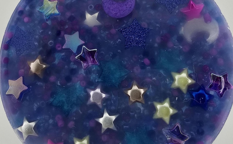 K&ouml;nnte beinhalten: Nahaufnahme eines blauen, glitzernden Harzornaments mit eingebetteten Sternen und einem Halbmond. Die Sterne sind in verschiedenen Farben gehalten, darunter Silber, Gold und Lila. Das Ornament hat ein himmlisches Thema.