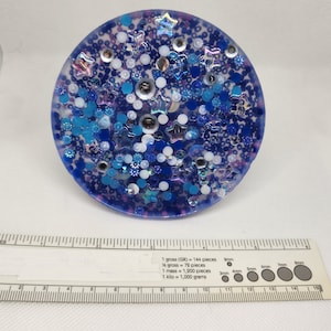 K&ouml;nnte beinhalten: Ein runder, blauer Harz-Untersetzer mit eingebetteten Glitzer, Sternen und Perlen in Blau-, Wei&szlig;- und Silbert&ouml;nen. Der Untersetzer hat einen Durchmesser von etwa 10 cm. Ein Lineal ist im Vordergrund.