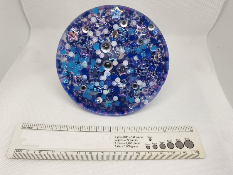 K&ouml;nnte beinhalten: Ein runder, blauer Harz-Untersetzer mit eingebetteten Glitzer, Sternen und Perlen in Blau-, Wei&szlig;- und Silbert&ouml;nen. Der Untersetzer hat einen Durchmesser von etwa 10 cm. Ein Lineal ist im Vordergrund.