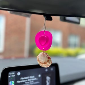 May include: A hot pink cowboy hat car charm with a seashell accent, hanging from a rearview mirror. The seashell is beige with reddish-brown markings. The car's infotainment screen is visible in the foreground.