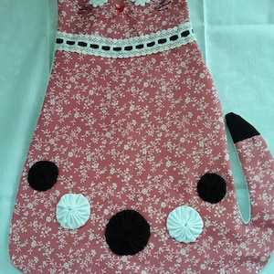May include: A cat-shaped fabric wall hanging in dusty rose with a white floral print. It features black fabric ears and tail tip, white daisy-shaped eyes, a red nose, and a white lace collar with black ribbon. Black and white fabric yo-yos decorate the body.