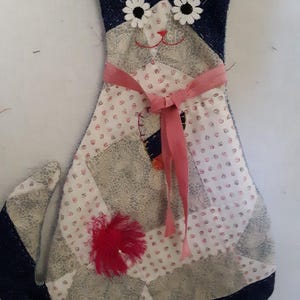 May include: A handmade cat-shaped decorative item. The cat is made of patchwork fabric in shades of blue, white, and gray. It features white daisy-like eyes, a pink ribbon, and a red fluffy embellishment. The item is likely a decorative pillow or wall hanging.