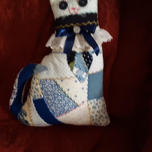 May include: A white patchwork cat pillow with blue and white fabric squares. The cat has black eyes, a black nose, and a blue bow around its neck.