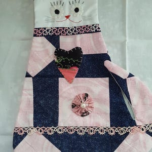 May include: A fabric cat-shaped wall hanging featuring a patchwork design in navy blue and light pink. The cat has embroidered white daisy eyes, a red nose, and whiskers. It is decorated with a black heart applique, a pink fabric flower, and lace.