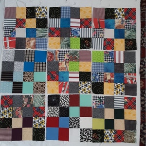 May include: A patchwork quilt top in progress, featuring a grid of colorful fabric squares. The squares display a variety of patterns, including stripes, florals, plaids, and geometric designs. The color palette includes red, blue, yellow, green, and black.