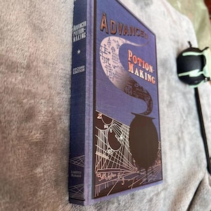 Puede incluir: Un libro de tapa dura azul titulado "Advanced Potion-Making" con una etiqueta de segunda edici&oacute;n. La portada presenta un caldero negro con vapor que sale de &eacute;l, rodeado de una ilustraci&oacute;n detallada de un patr&oacute;n de telara&ntilde;a.