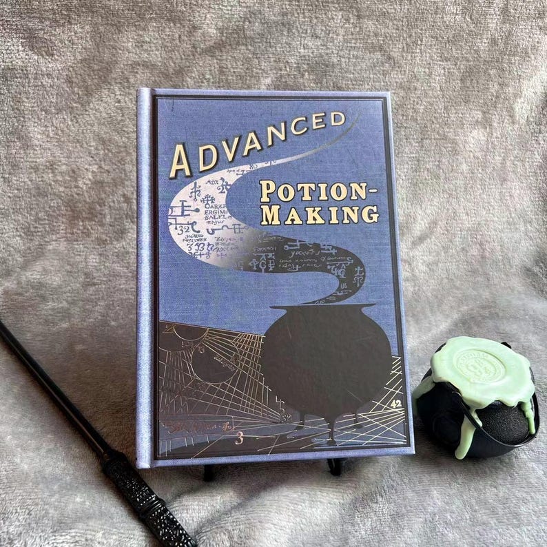 Puede incluir: Un libro azul con el t&iacute;tulo "Advanced Potion-Making" en la portada. El libro presenta un caldero negro con vapor que sale de &eacute;l. Un peque&ntilde;o caldero verde con un l&iacute;quido verde est&aacute; en primer plano.