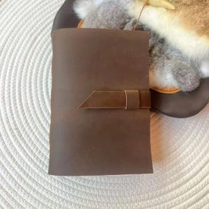 May include: A brown leather journal with a unique angled flap closure. The journal is closed, with a simple, elegant design. The leather has a rich, dark brown color, suggesting quality craftsmanship. The journal is likely for writing or sketching.