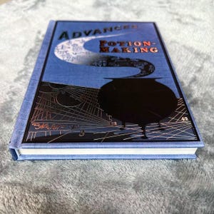 Puede incluir: Un libro de tapa dura azul con el t&iacute;tulo "Advanced Potion-Making" en letras doradas. La portada presenta un caldero negro con un dise&ntilde;o plateado en espiral.