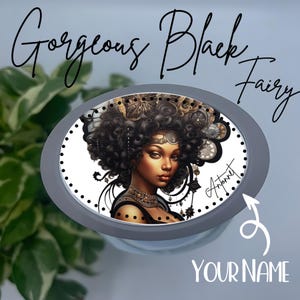 May include: A round, gray phone grip with a white background featuring a black woman with a large afro, wearing a gold crown and necklace. The text "Antonet" is written in cursive on the right side of the image. The words "Gorgeous Black Fairy" are written in black cursive at the top of the image.