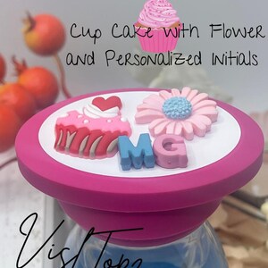 May include: Pink and white cupcake with pink frosting and sprinkles, a pink flower with a blue center, and pink, blue, and white letters spelling "MG" on a pink silicone bottle stopper. The text "Cup Cake with Flower and Personalized Initials" is written above the bottle stopper.
