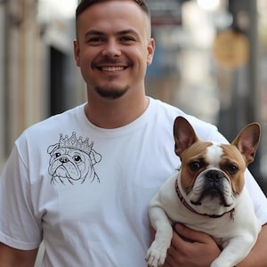 Puede incluir: Una camiseta blanca de manga corta con un gráfico de líneas negras de un bulldog con corona en la parte superior del pecho. La prenda está confeccionada en tela blanca lisa y cuenta con un cuello redondo estándar de aspecto sencillo.