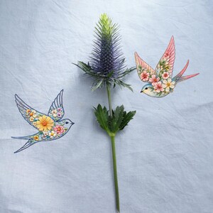 Puede incluir: Una composición con una flor de cardo con una cabeza púrpura y espinosa y dos golondrinas decorativas. Las golondrinas están adornadas con motivos florales en rosa, amarillo y blanco, sobre un fondo azul claro.