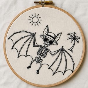 Puede incluir: Un bastidor de bordado circular de madera con tela blanca y bordado en hilo negro. El diseño muestra un murciélago esqueleto con sombrero de sol y gafas de sol, flanqueado por un icono de sol a la izquierda y una palmera a la derecha.