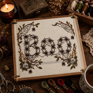 Puede incluir: Un bastidor de bordado cuadrado que contiene un panel de tela blanca con la palabra BOO bordada en hilo negro, formada por pequeñas arañas con detalles rojos. El diseño incluye telarañas, hojas verdes, dos calaveras y velas encendidas.