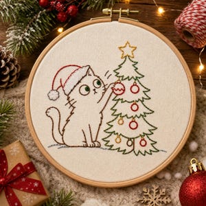 Puede incluir: Un bastidor de bordado circular de madera que contiene un panel de tela blanca con un diseño bordado a mano de un gato blanco con un gorro de Papá Noel rojo y blanco, decorando un árbol de Navidad verde con adornos circulares rojos y dorados.