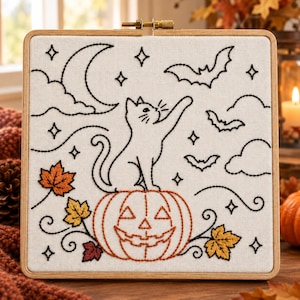 Puede incluir: Un bastidor de bordado cuadrado con tela blanca que presenta un dibujo lineal negro de un gato sentado sobre una calabaza, rodeado de murciélagos, nubes, estrellas y una luna creciente. La calabaza y las hojas otoñales están bordadas.
