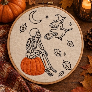 Puede incluir: Un bastidor de bordado redondo de madera con una tela beige que presenta un bordado en hilo negro de un esqueleto sentado sobre una calabaza naranja brillante, una bruja volando en escoba, una luna creciente, estrellas y hojas.