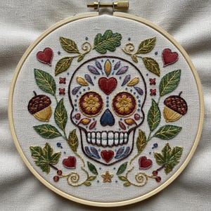 Puede incluir: Un bastidor de bordado circular de madera que contiene un panel de tela con un diseño de calavera de azúcar bordado a mano. La calavera está decorada con coloridos motivos florales, corazones y hojas, flanqueada por dos bellotas y bayas rojas.