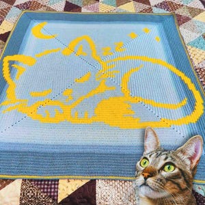 Puede incluir: Una manta de ganchillo azul con un diseño de gato amarillo. La manta presenta un efecto degradado y un borde amarillo. El diseño del gato incluye una luna creciente y estrellas. Mide aproximadamente 120 cm x 120 cm.