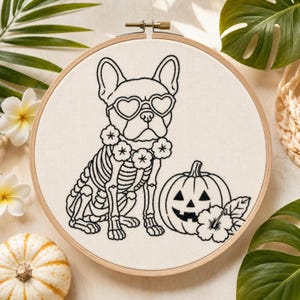 Puede incluir: Un bastidor de bordado circular con un diseño de líneas negras sobre tela blanca. El dibujo muestra un esqueleto de bulldog francés con gafas de sol en forma de corazón y un collar de flores, junto a una calabaza y una flor de hibisco.