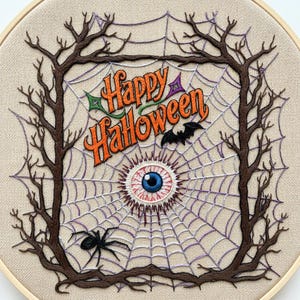 Puede incluir: Un bastidor de bordado circular con fondo de tela beige y un diseño cosido de ramas de árbol oscuras que forman un marco cuadrado. En el interior, hay una telaraña morada, una araña negra, un murciélago negro y un ojo azul bordados. El texto Happy Halloween está bordado en naranja.